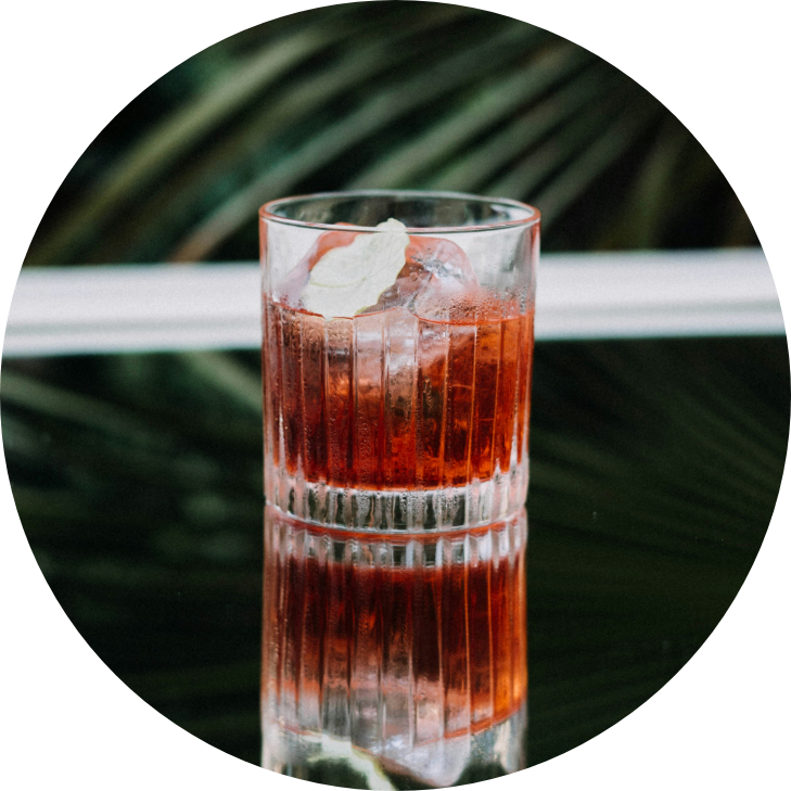 A glass of a red-colored beverage with ice and a lime wedge, placed on a reflective surface with a blurred green background.