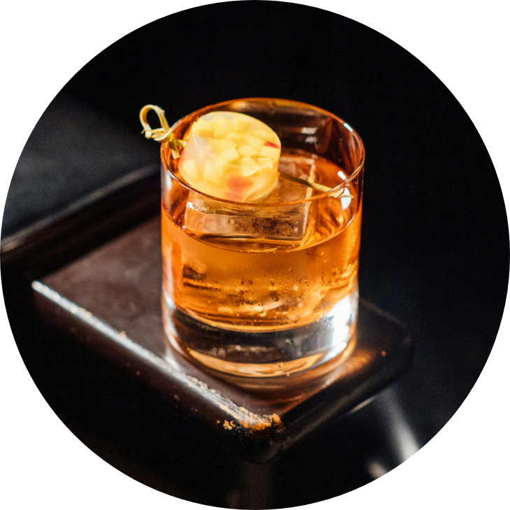 A glass of whiskey on a black surface with a sugar cube on a cocktail pick garnished with a piece of lemon peel.