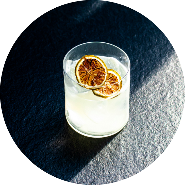 A glass of clear lemonade with ice and dried lemon slices garnishing the top, placed on a dark textured surface.