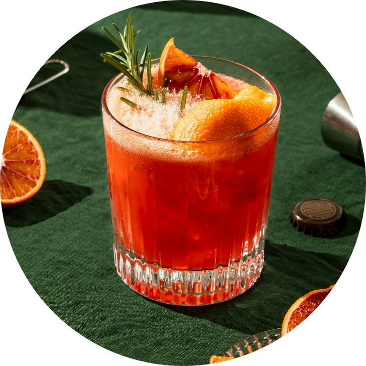 A glass of red cocktail garnished with orange and blood orange slices and a sprig of rosemary, on a green tablecloth with additional blood orange slices and a bottle cap nearby.