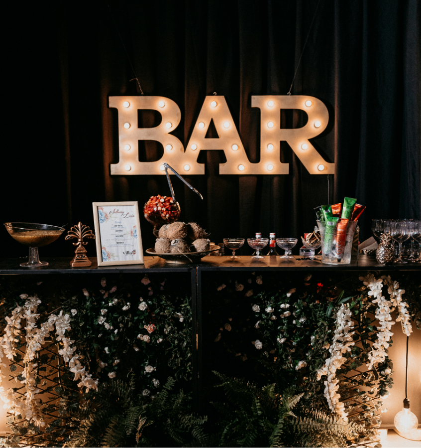 Decorative bar setup with large illuminated 'BAR' sign, various bowls and bottles, and floral decorations beneath.