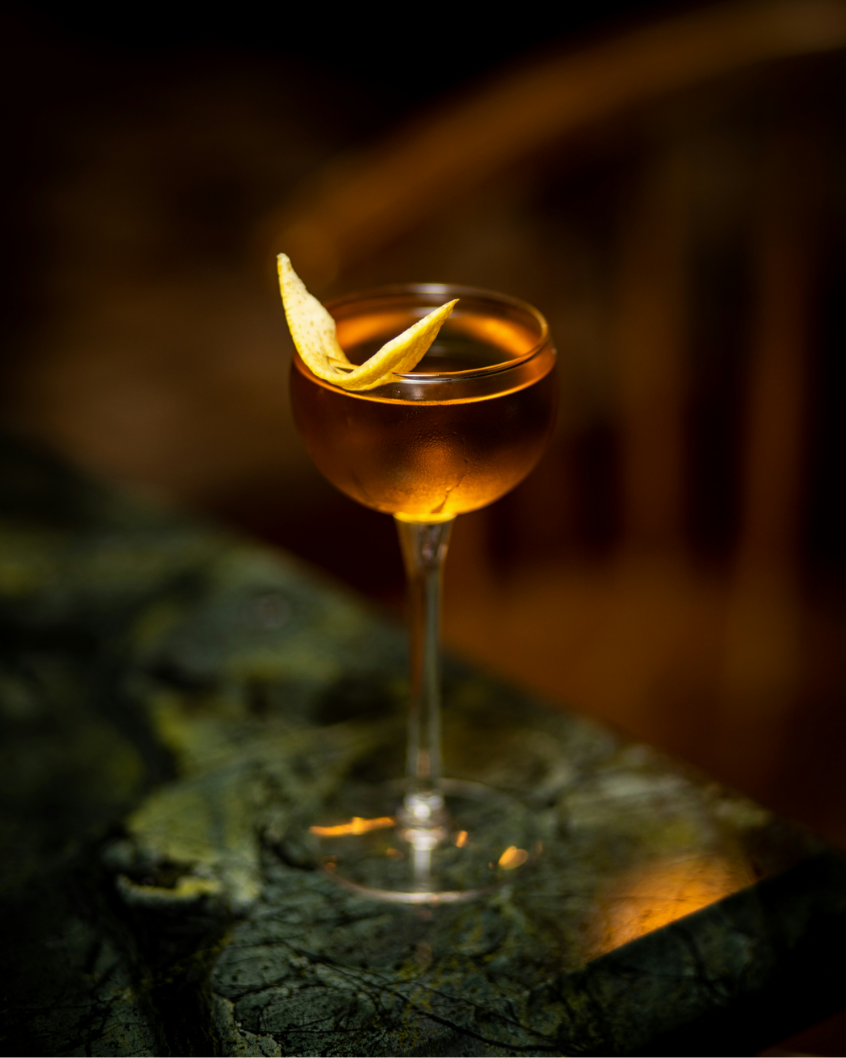 A cocktail in a coupe glass garnished with a lemon peel on a dark, textured surface.