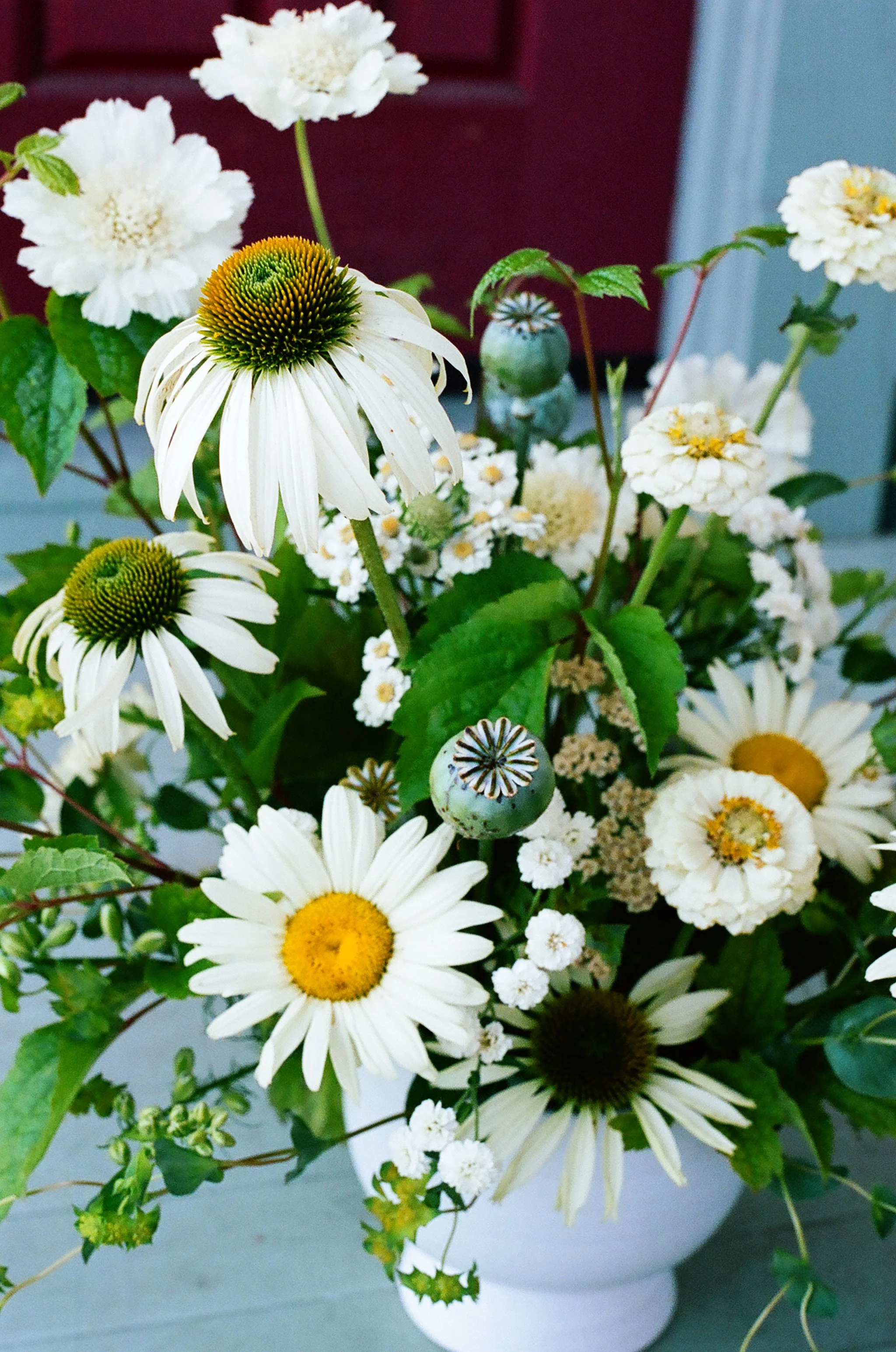 A bouquet of white daisies with yellow centers, white yarrow flowers, and some green leaves in a white vase.