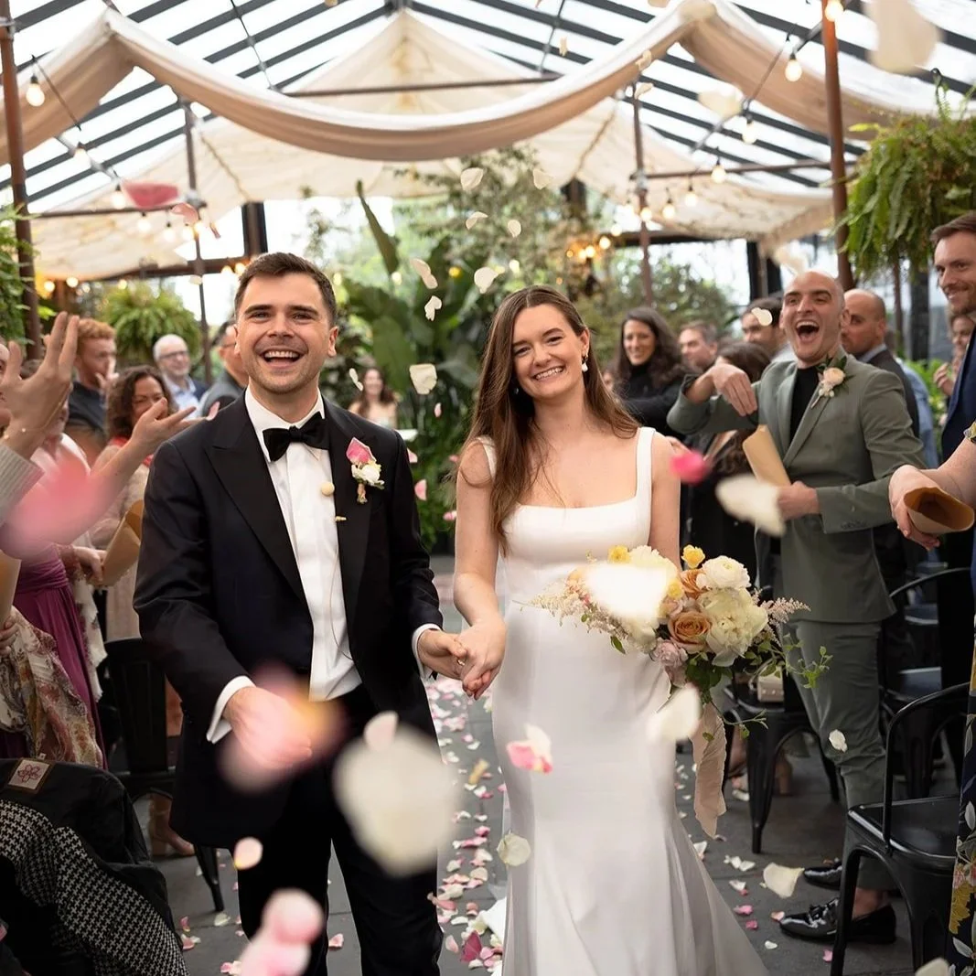 A bride and groom holding hands and smiling as they walk down the aisle during their wedding celebration, surrounded by happy guests throwing flower petals in an indoor venue decorated with string lights and greenery.