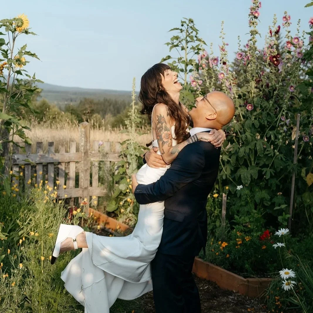 A joyful woman in a white dress with tattoos hugging and laughing with a bald man in a black suit in a garden with flowers and greenery.