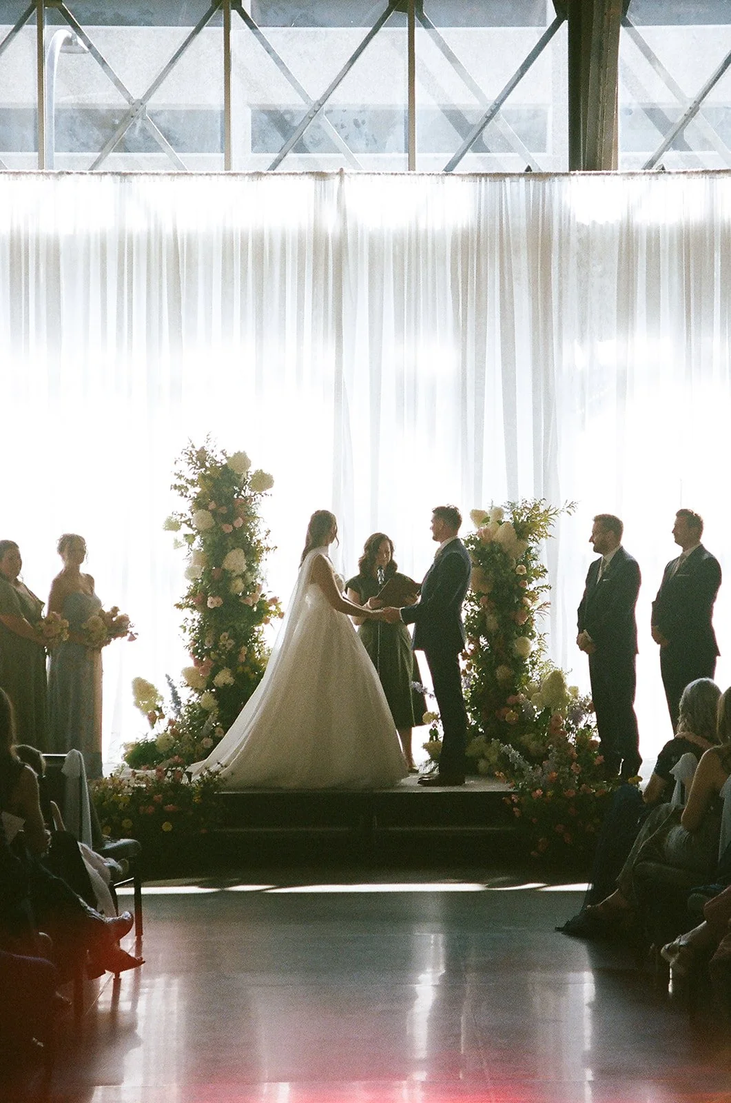 A wedding ceremony taking place indoors with a bride and groom exchanging vows at the altar, surrounded by floral arrangements, with wedding parties standing on either side and guests seated in the foreground.