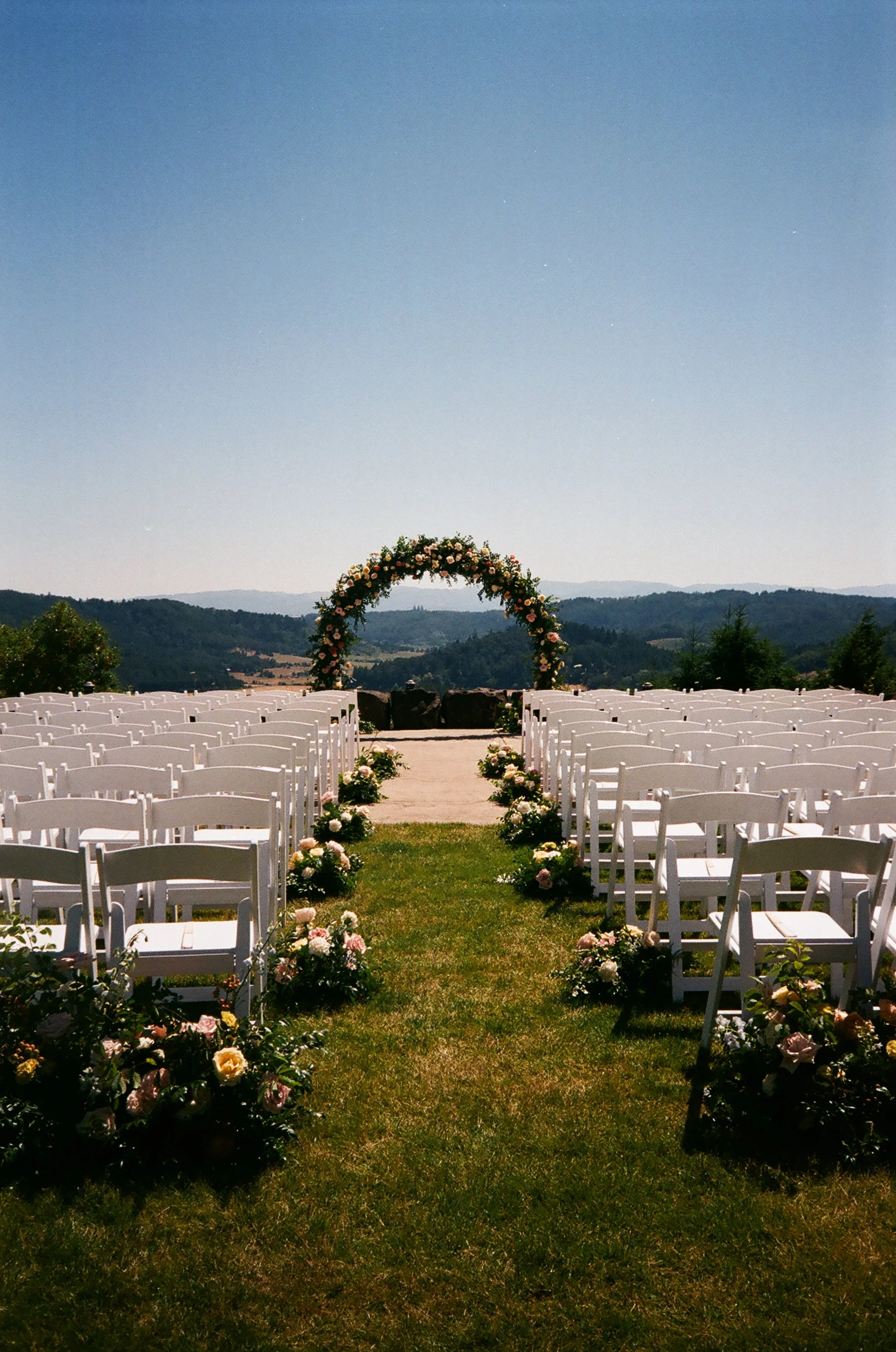 Outdoor wedding ceremony with hilltop mountain view, white folding chairs, grass aisle lined with floral arrangements, and a floral arch in peach, blush, and yellow blooms, floral design by Faded Era Floral.