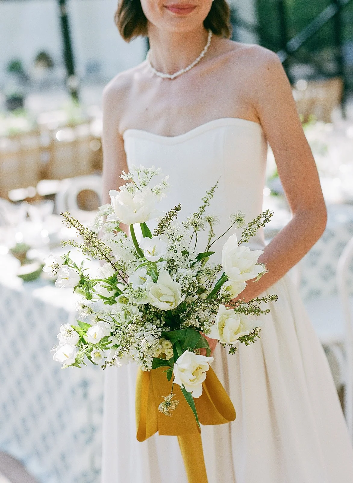 A woman dressed in a white strapless gown holds a bouquet of white flowers, decorated with a yellow ribbon, at a wedding reception.