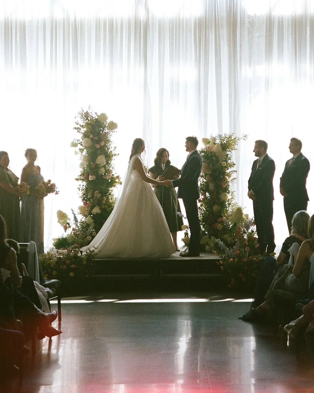 A wedding ceremony with the bride and groom holding hands and facing each other, surrounded by bridesmaids and groomsmen, with floral arrangements and backlit curtains.
