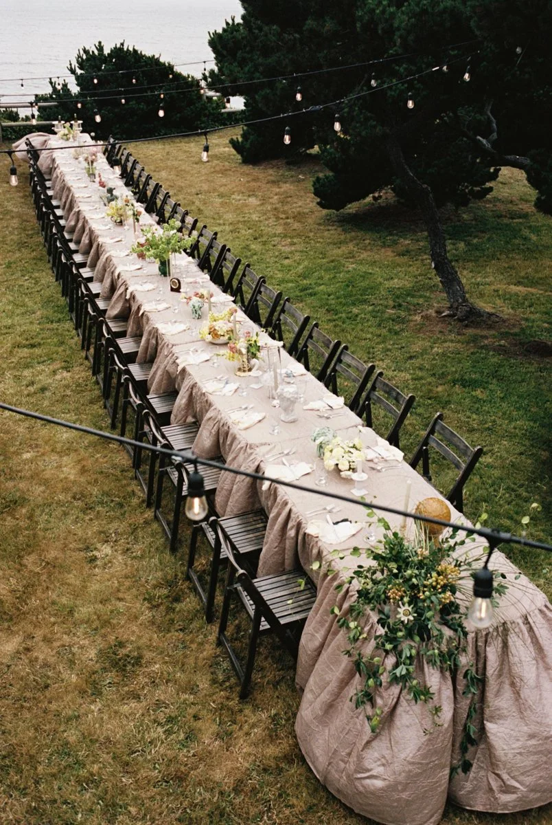 An outdoor dining table set for a celebration, decorated with flowers and candles, with string lights overhead and trees in the background.