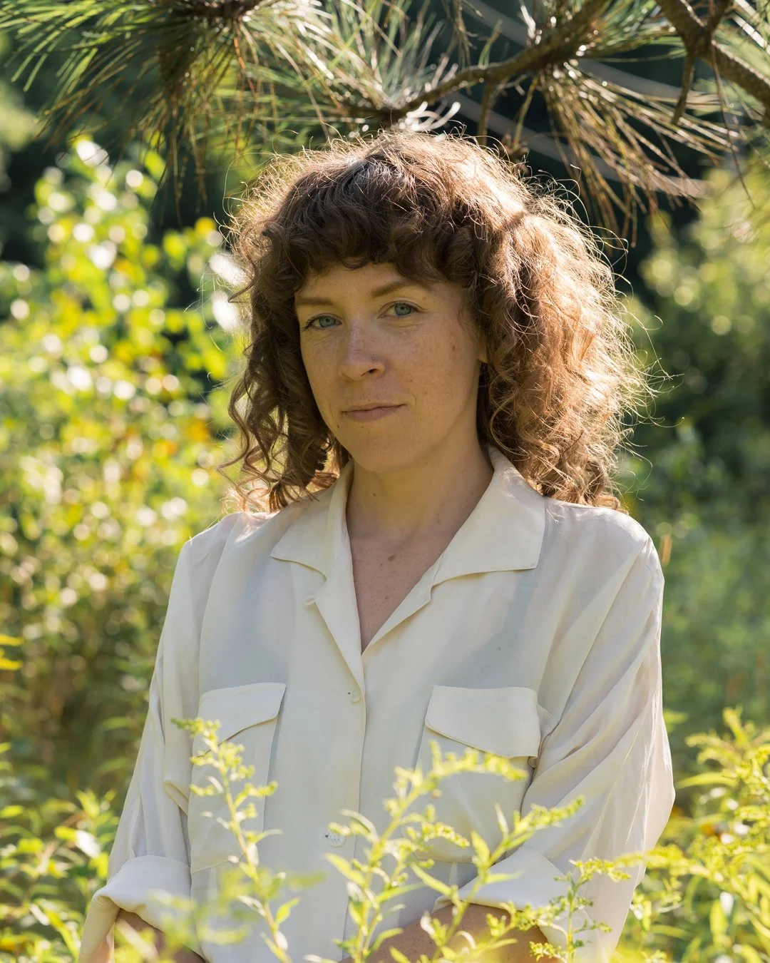 A woman with curly brown hair and blue eyes standing outdoors among green plants and trees, wearing a cream-colored blouse.