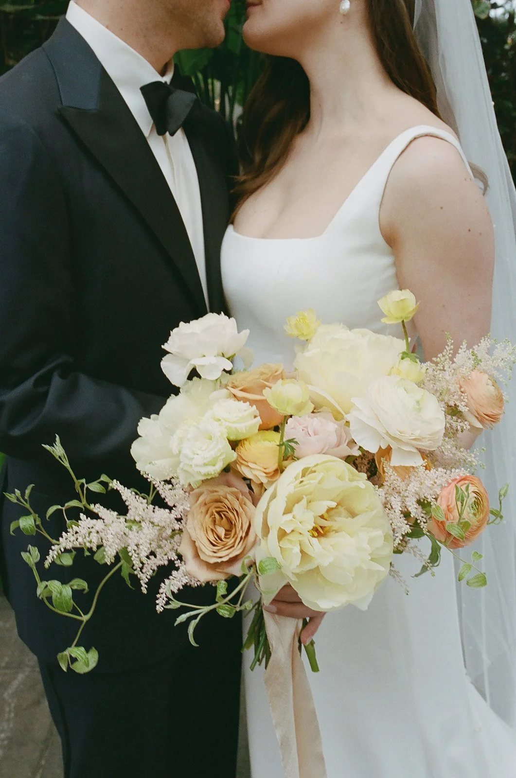 A bride and groom are facing each other, holding a bouquet of flowers at their wedding. The bride is wearing a white dress with spaghetti straps and a pearl earring. The groom is dressed in a black tuxedo with a black bow tie.