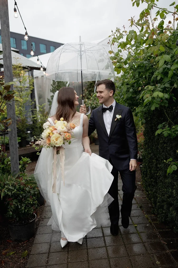 A bride and groom walking outdoors on a rainy day, sharing an umbrella. The bride is holding a bouquet of flowers and lifting her dress slightly, smiling at the groom. The groom is dressed in a black tuxedo with a bow tie, smiling back at the bride. Green foliage surrounds them.