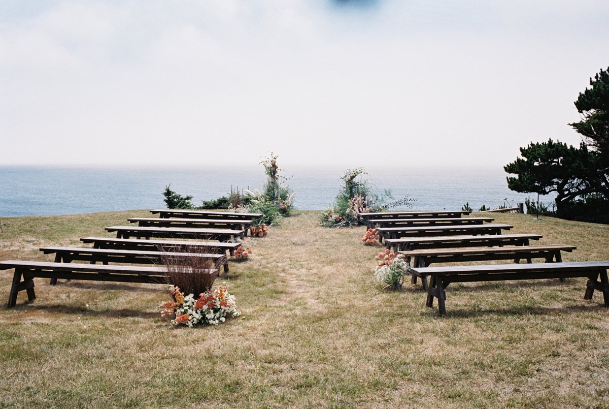 Outdoor wedding ceremony setup with wooden benches arranged facing an ocean view, decorated with floral arrangements on either side, during daytime.
