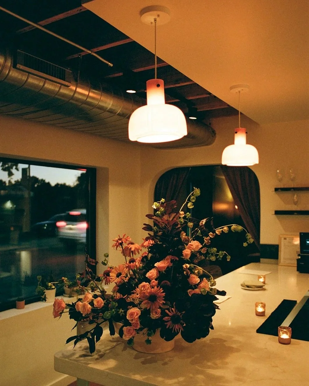 Interior of a dimly lit cafe or restaurant featuring two hanging ceiling lights, a large flower arrangement on the counter, and a window showing blurred cars outside during dusk.