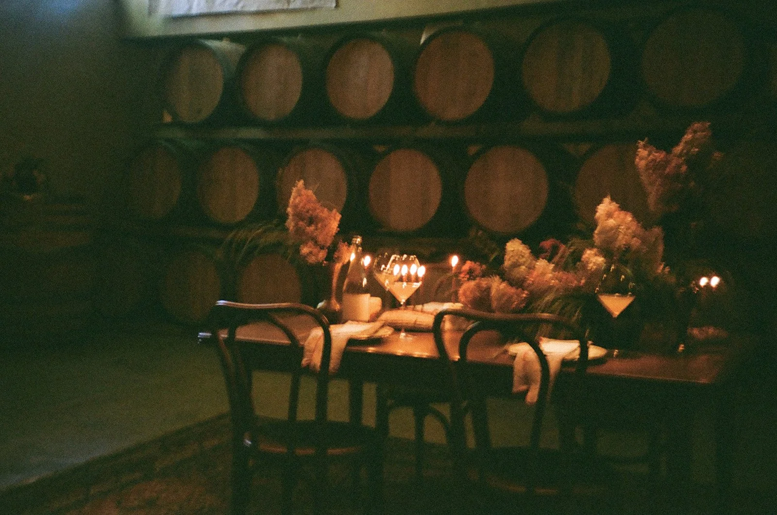 A dimly lit vintage dining setup with a table decorated with flowers and candles against a wall of large wooden barrels.
