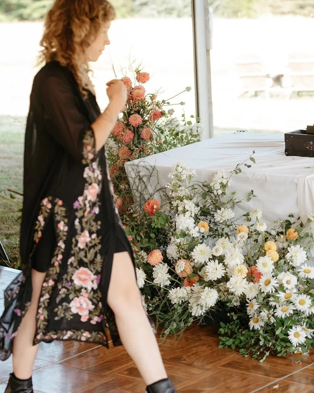 A woman with red hair in a black floral dress is adjusting flowers near a decorated table with white and peach flowers, set outdoors during daylight.