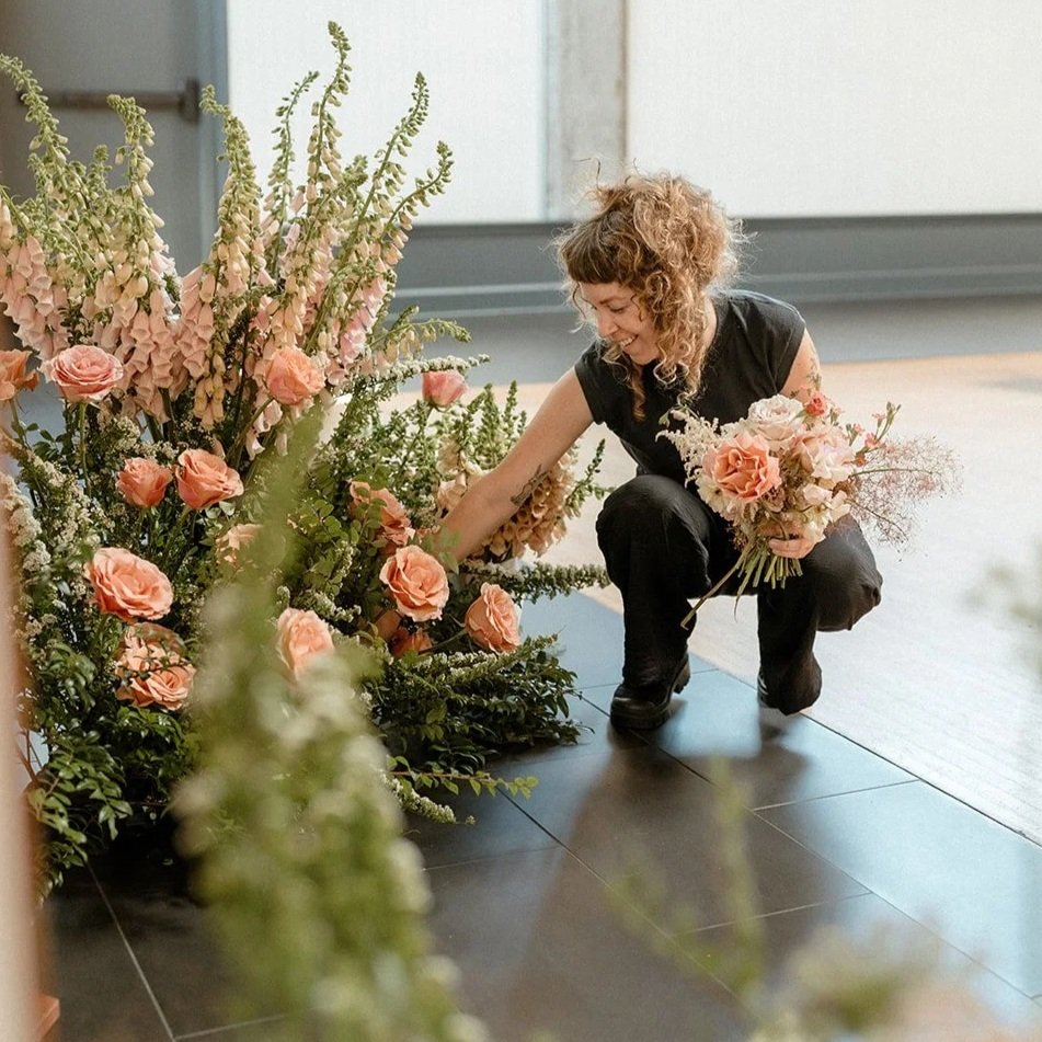 A woman arranging a floral display with pink and white flowers, smiling as she crouches on a black tiled floor inside a room.