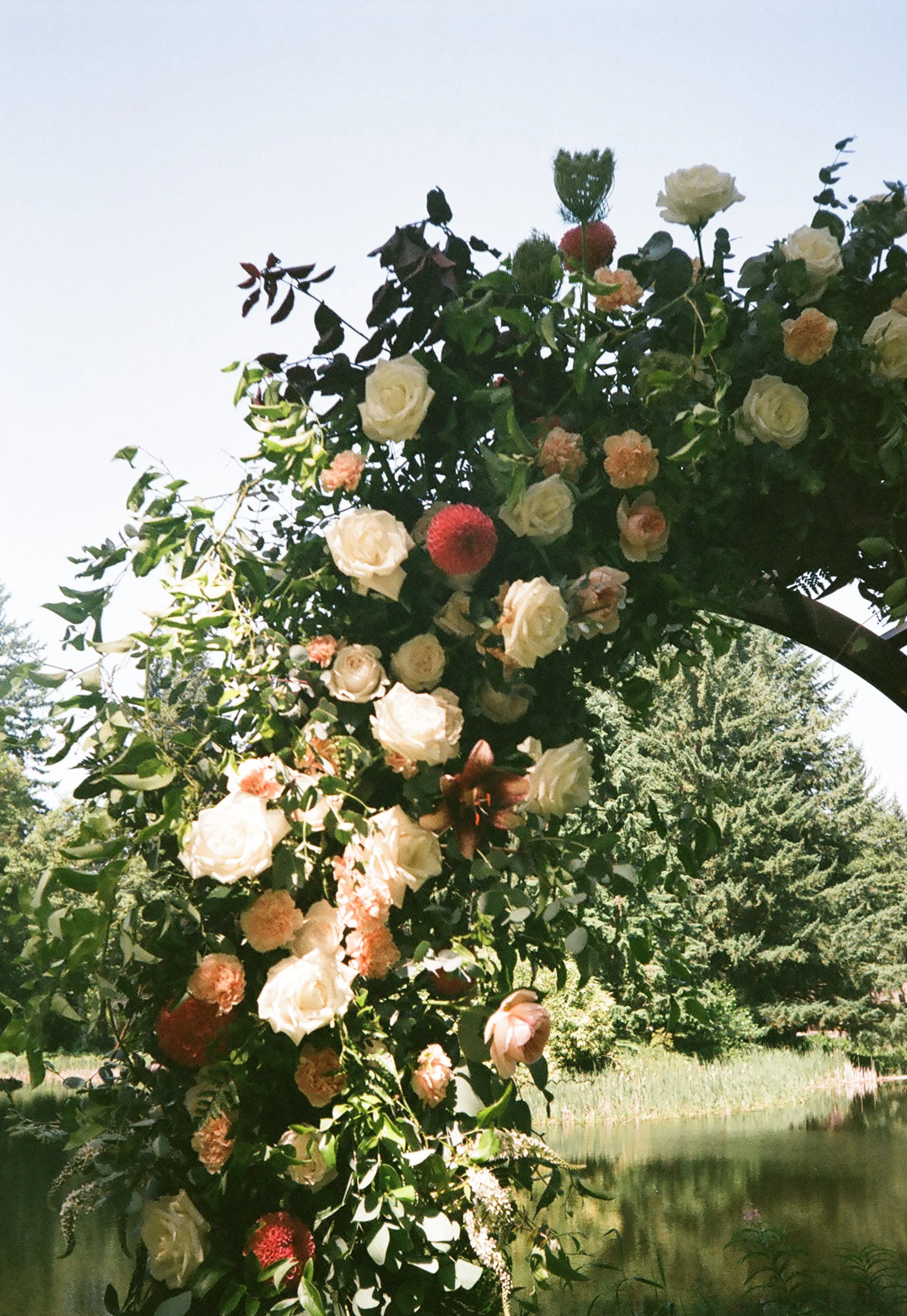 A floral arrangement hanging above a pond, featuring white roses, pink and red flowers, with lush green foliage, in a natural outdoor setting.