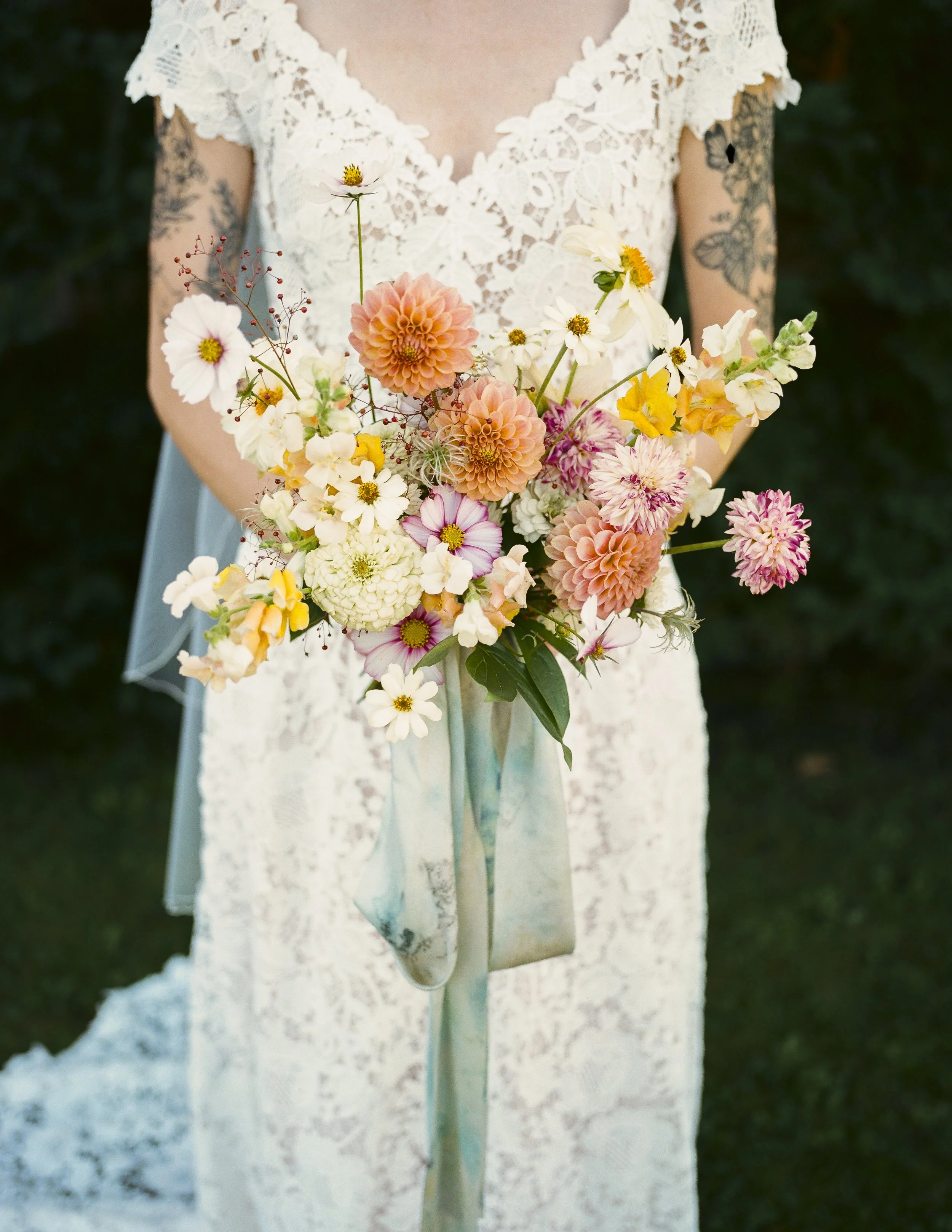A person wearing a white lace dress holding a bouquet of various colorful flowers, including dahlias and daisies, outdoors with dark green foliage in the background.