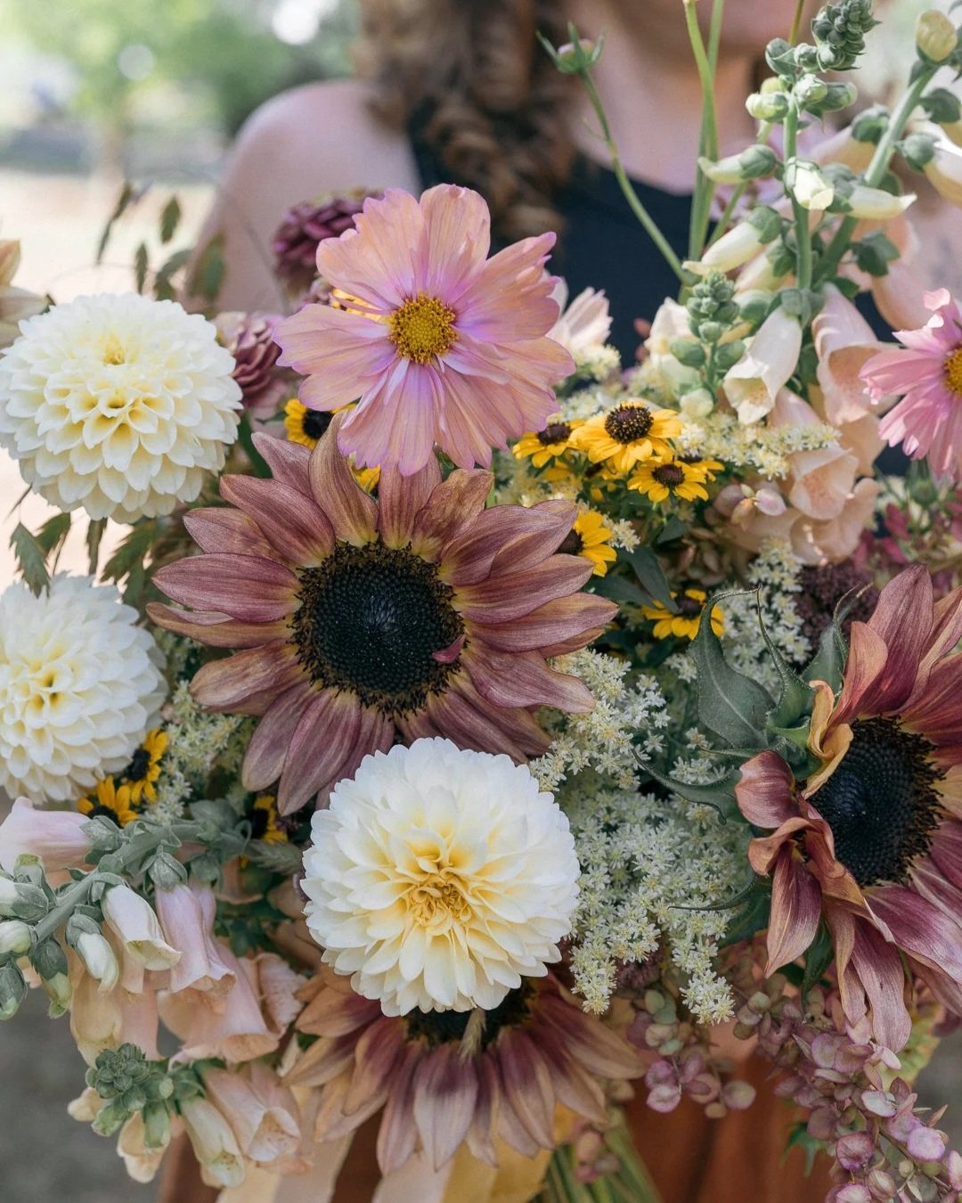 A person holding a large bouquet of colorful flowers including sunflowers, dahlias, and various small yellow, white, and pink blossoms.