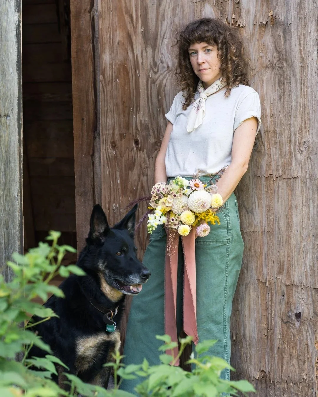 A woman holding a bouquet of flowers, standing next to a black dog, in front of a rustic wooden wall.