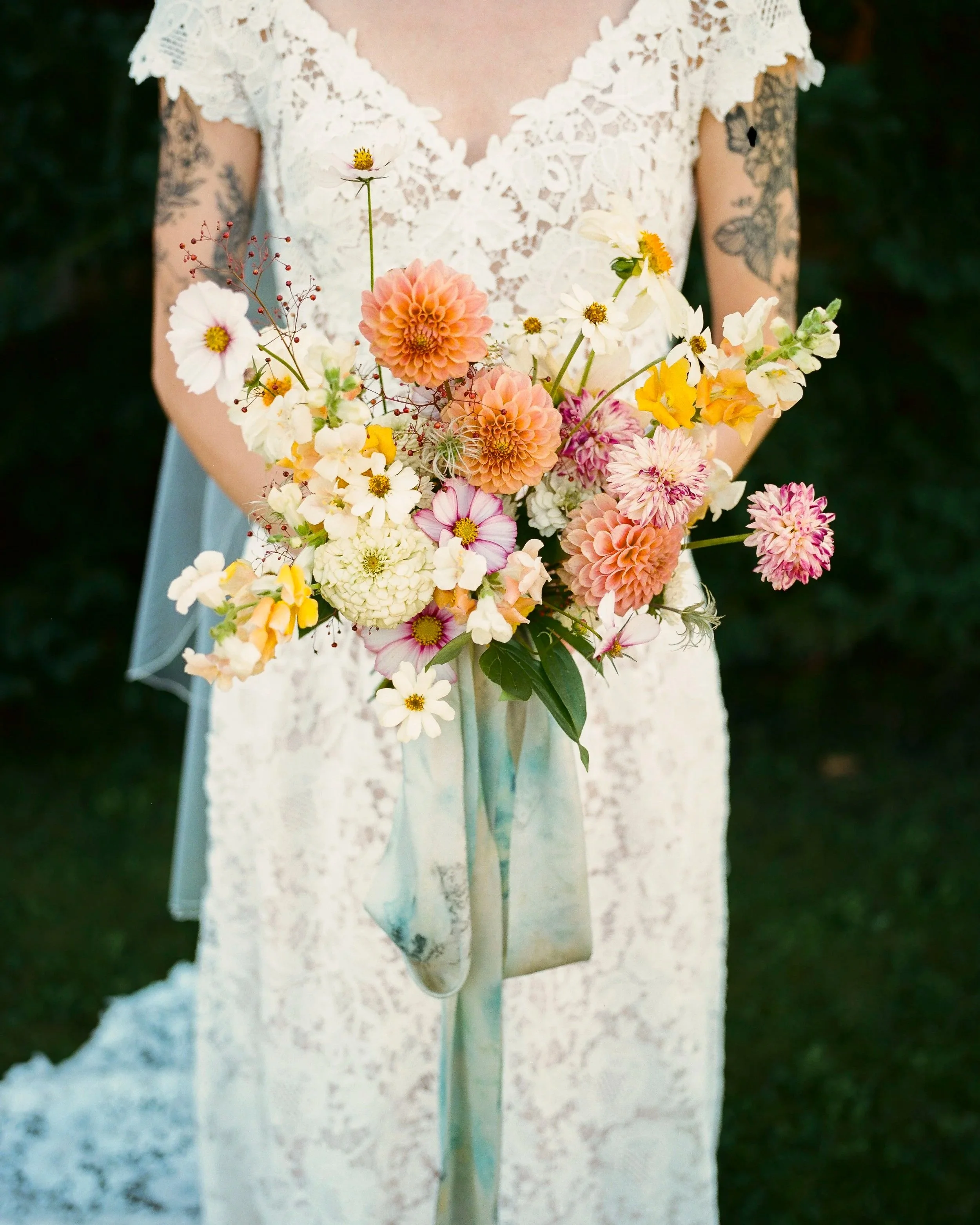 Close-up of a person wearing a lace dress holding a colorful bouquet of flowers, showing only the torso and arms, with tattoos on both arms, against a dark green background.