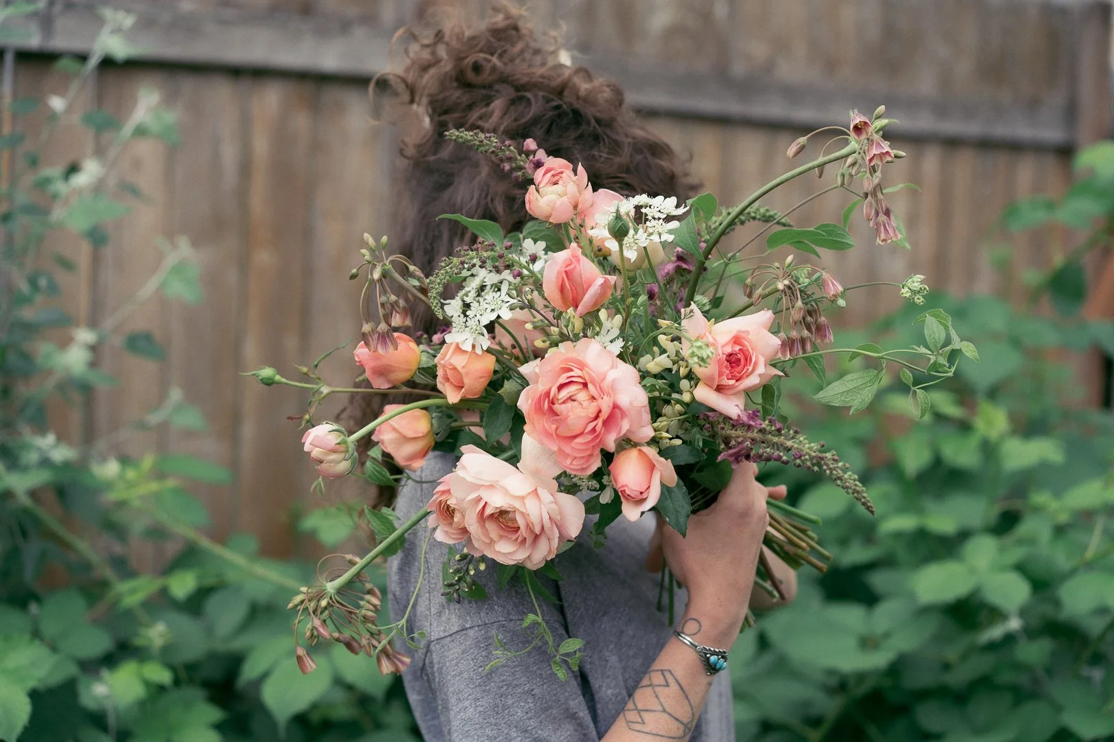 A woman holding a large bouquet of pink and white flowers in front of her face, standing outdoors in front of a wooden fence and green plants.
