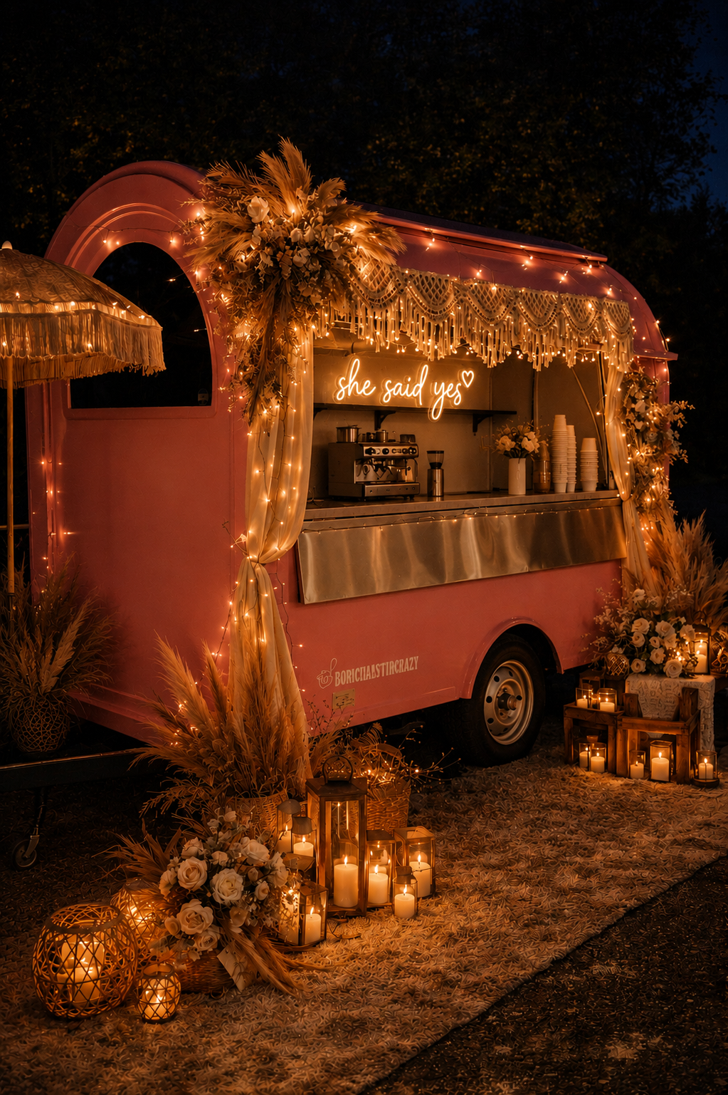 A decorated pink food truck at night with warm string lights, floral arrangements, candles, and a neon sign saying 'she said yes' for a celebration or wedding event.
