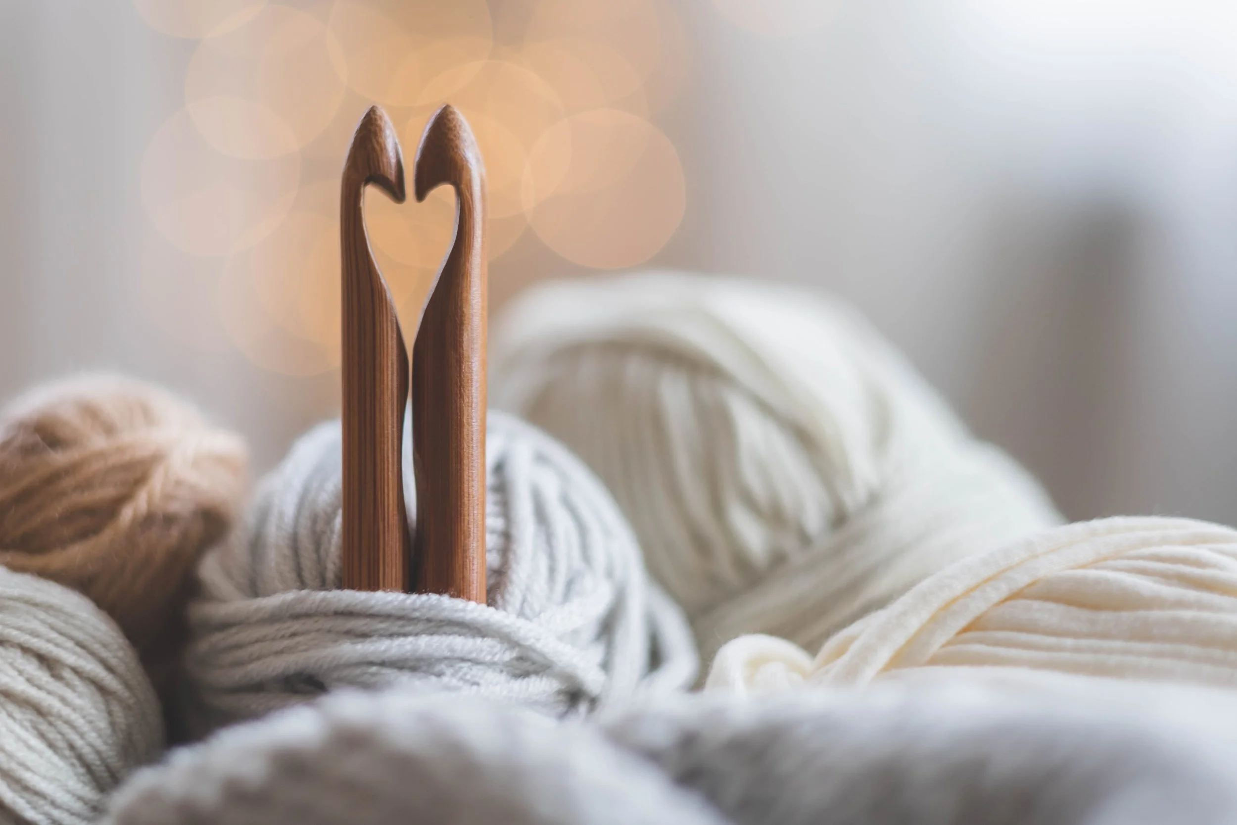 Two wooden knitting needles with a heart shape formed between their tips, resting on bundles of cream and beige yarn.