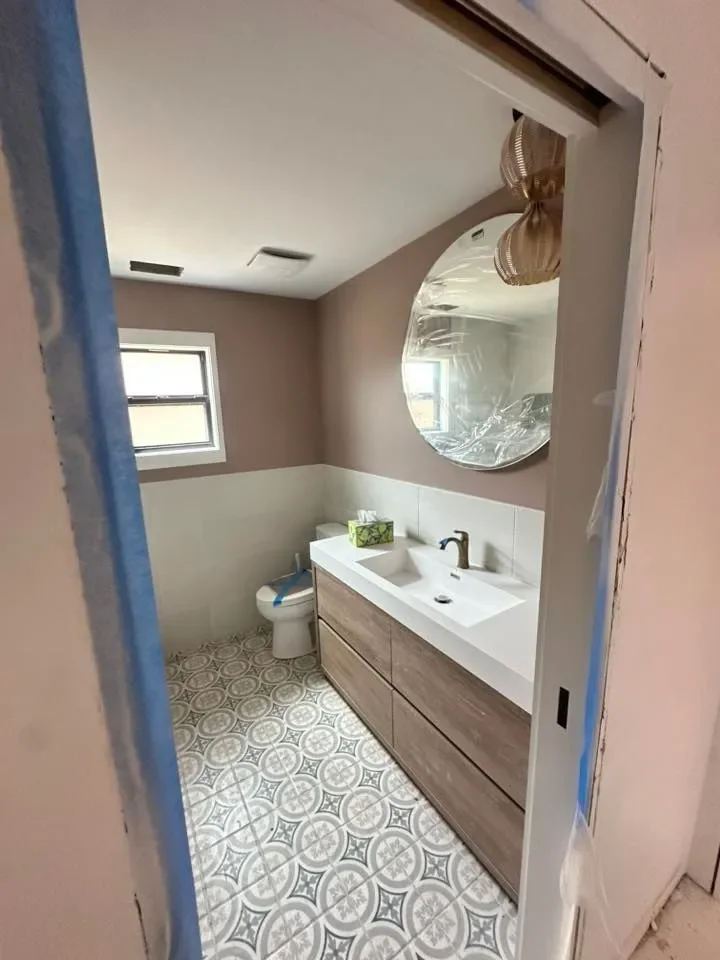 View of a modern bathroom with a wooden vanity, white sink, and gold faucet. A round mirror and a small window above the toilet are visible. The bathroom features patterned tile flooring and beige walls.