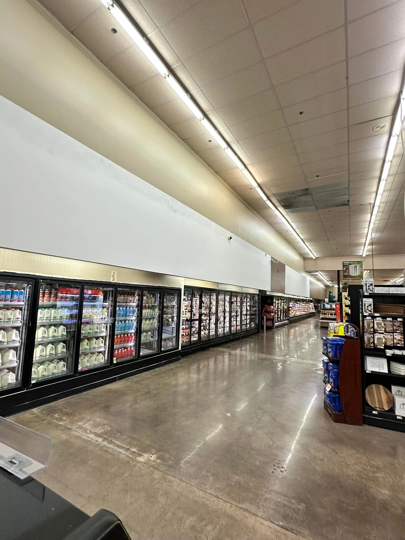 A grocery store aisle with refrigerated section on the left, shelves stocked with dairy products, and shoppers in the distance.
