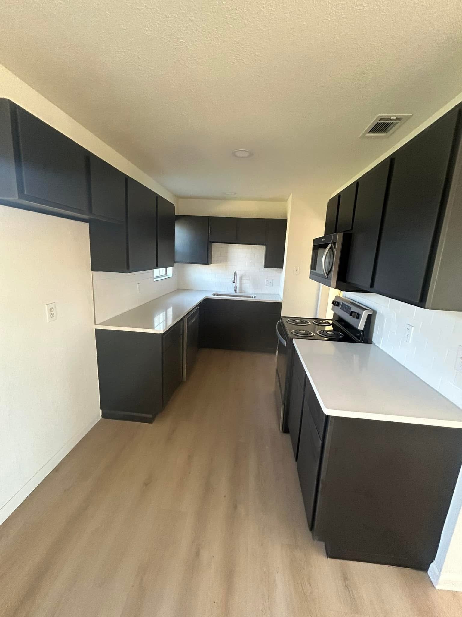 A modern kitchen with black cabinets, white countertops, a stainless steel microwave, stove, and a sink under a window. Beige flooring and white walls with minimal decor.