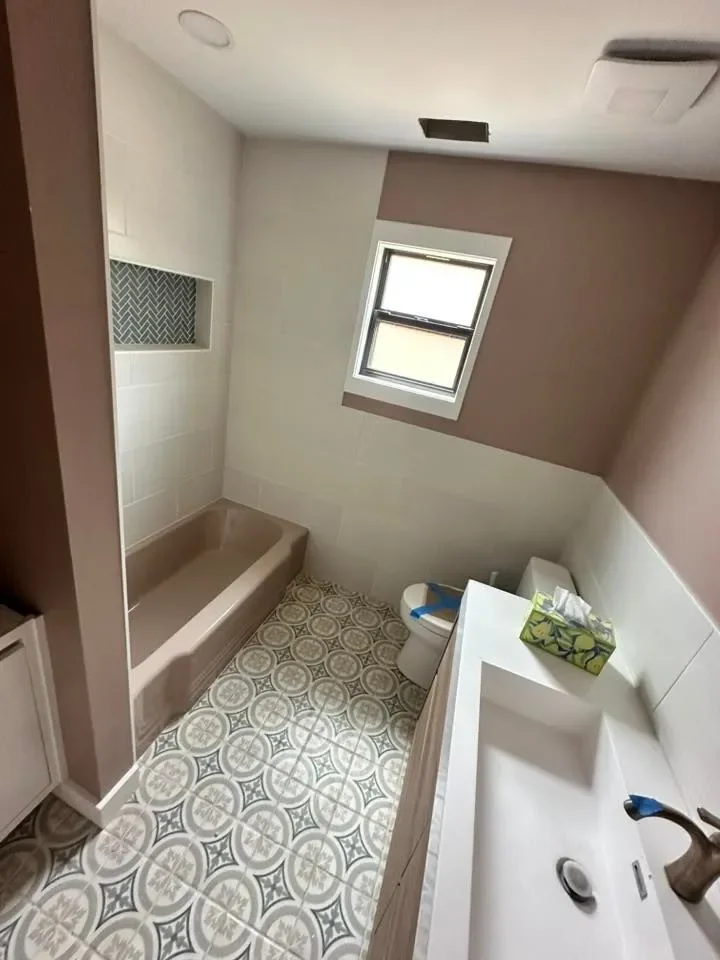A small bathroom with patterned floor tiles, a window, a bathtub, a toilet, and a sink with a tissue box on it.