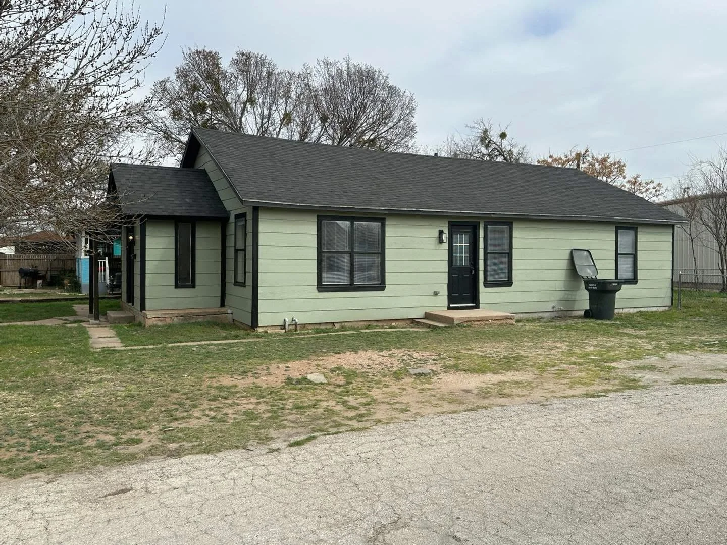 A single-story house with light green siding and black trim, featuring multiple windows and a black door. A garbage bin is outside, and there's a small porch step. The front yard has patchy grass and gravel, with a few steps leading to the door and a