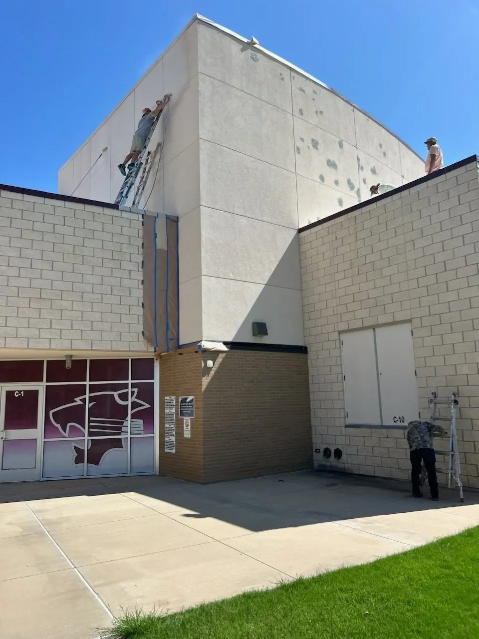 Two workers are painting the exterior wall of a building. One is on a ladder, painting higher on the wall, while the other is standing on the ground near a ladder, preparing to work. The building has a large glass window with an eagle logo and signs 
