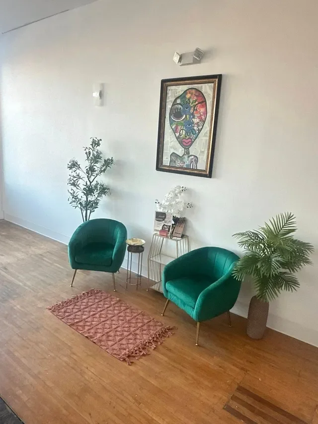 A modern sitting area with two green velvet armchairs, a pink textured rug, two large potted plants, a small side table with a decorative object, a magazine rack, and a colorful abstract portrait on the wall.