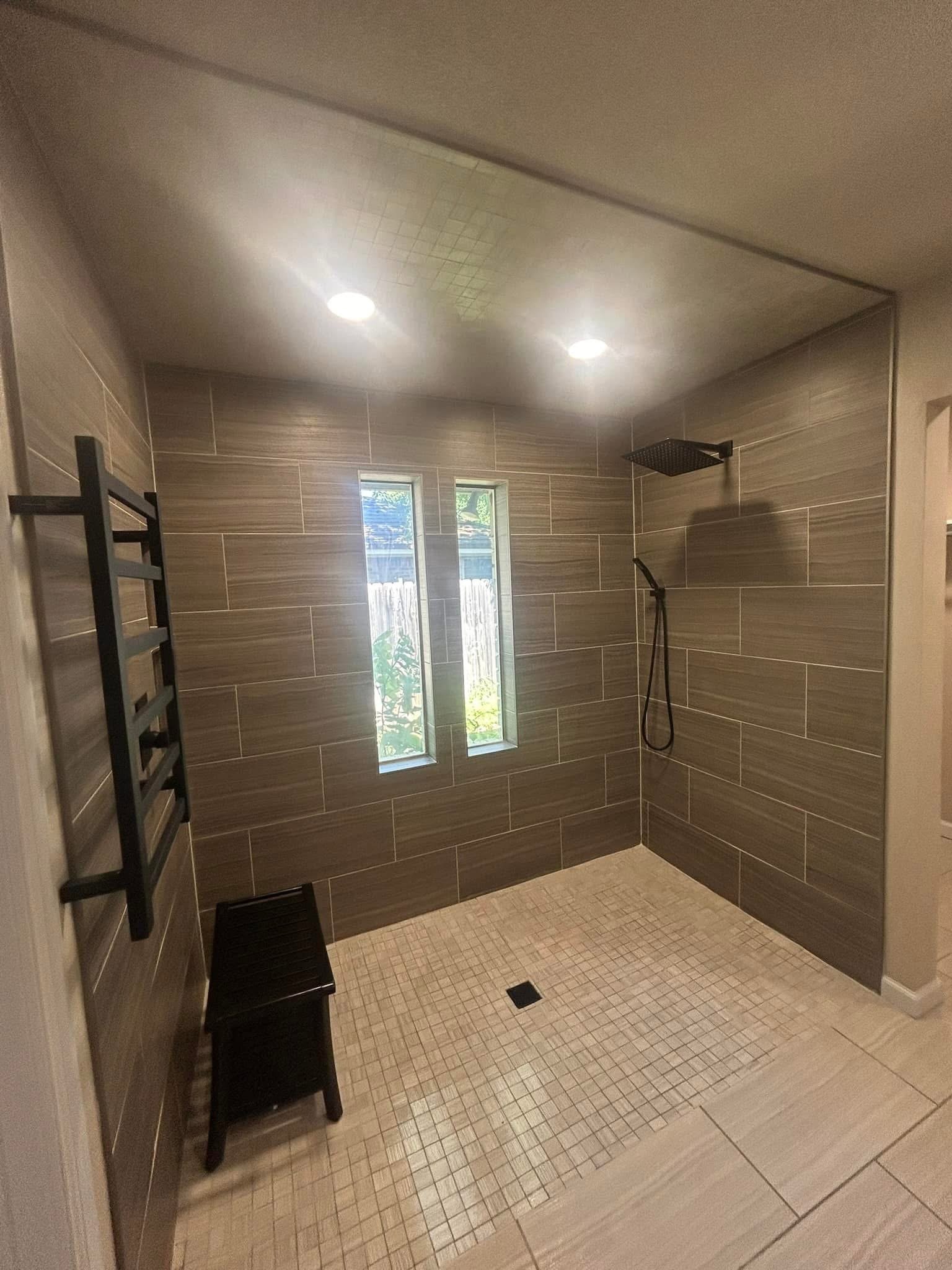A modern walk-in shower with beige tiled walls, a rainfall showerhead, and a window showing greenery outside. There is a small black stool and a black towel rack on the left.