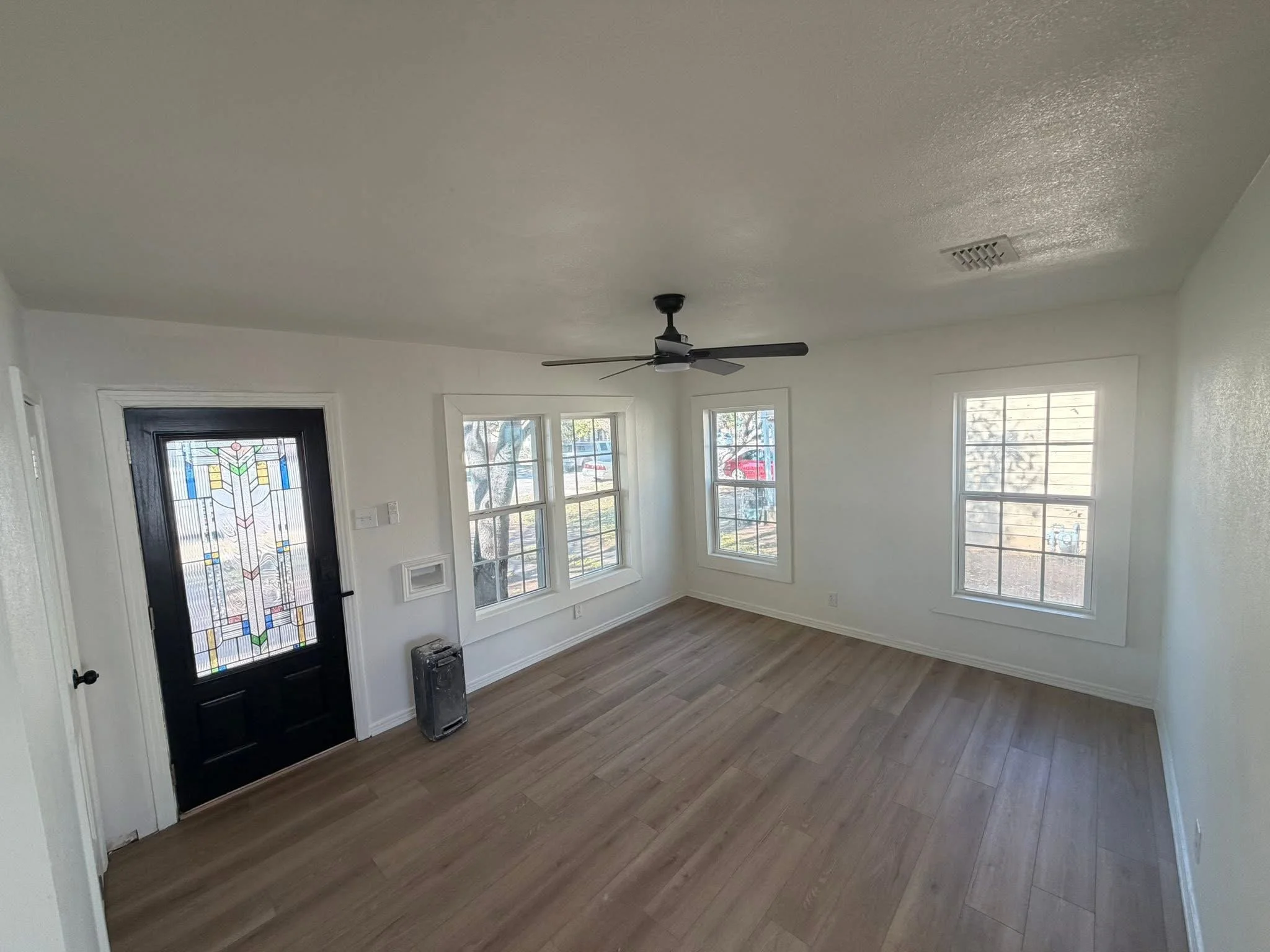 Interior view of a bright, empty living room with white walls, multiple windows, wood flooring, a black front door with stained glass, ceiling fan, and a small heater near the window.