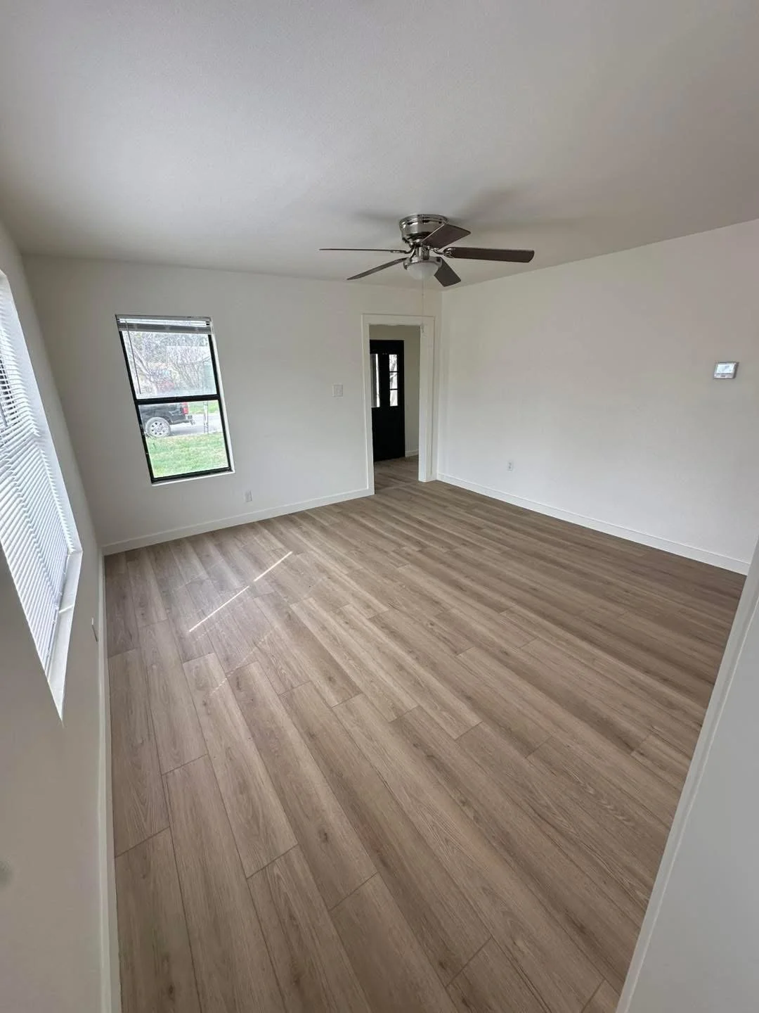 Empty living room with white walls, wood laminate flooring, a ceiling fan, and two windows.