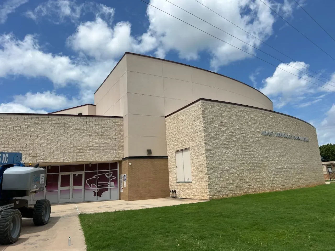 Exterior of a school building with a curved stone wall, glass doors with school logos, and a grassy area in front.