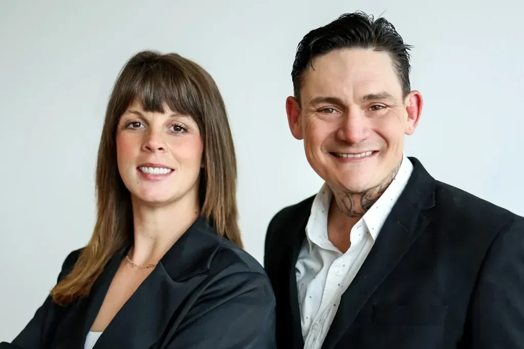 A man and woman wearing business attire smiling at the camera against a plain background.