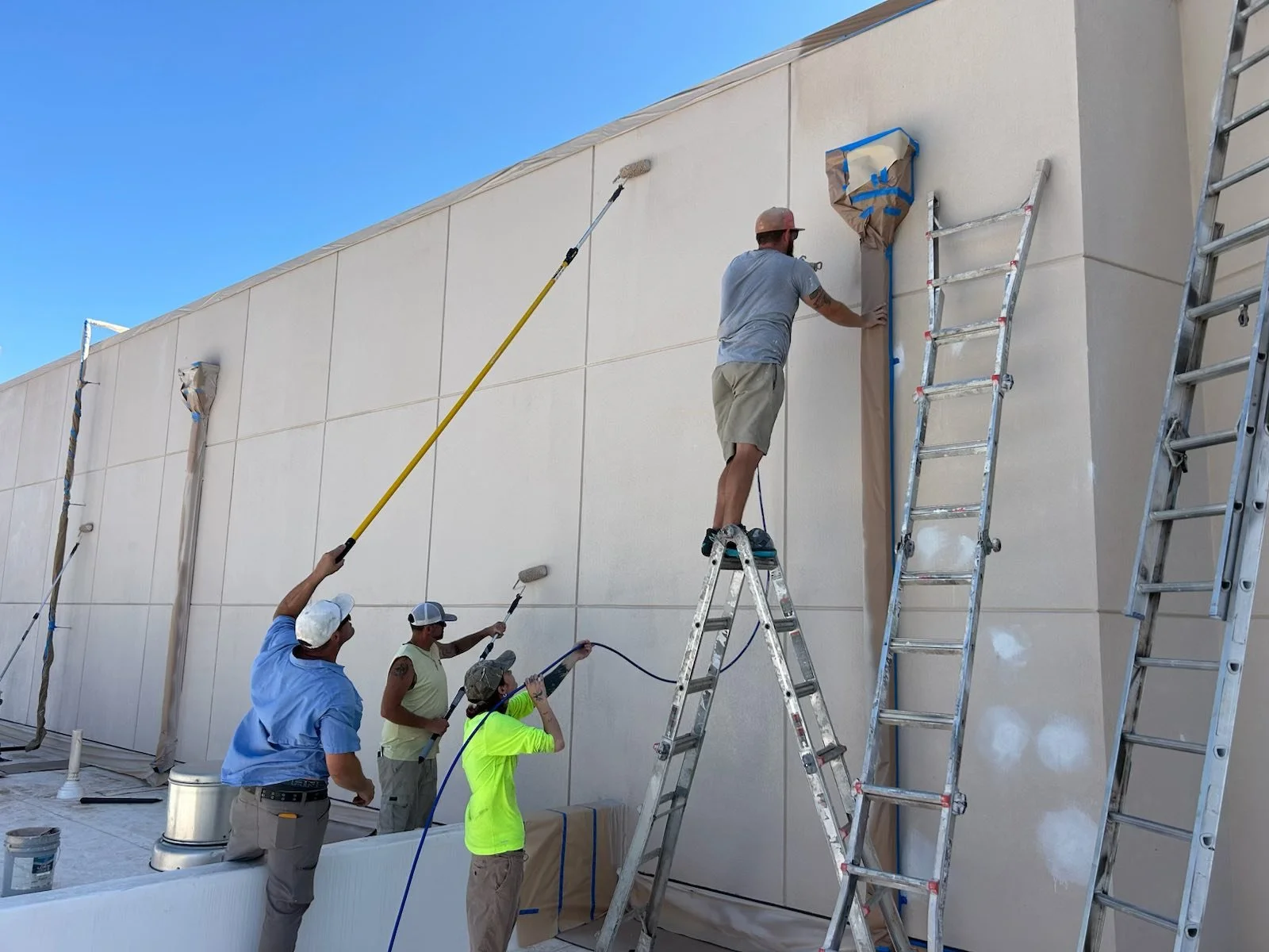 Group of workers painting the exterior wall of a building using paint rollers, with ladders and scaffolding nearby under a clear blue sky.