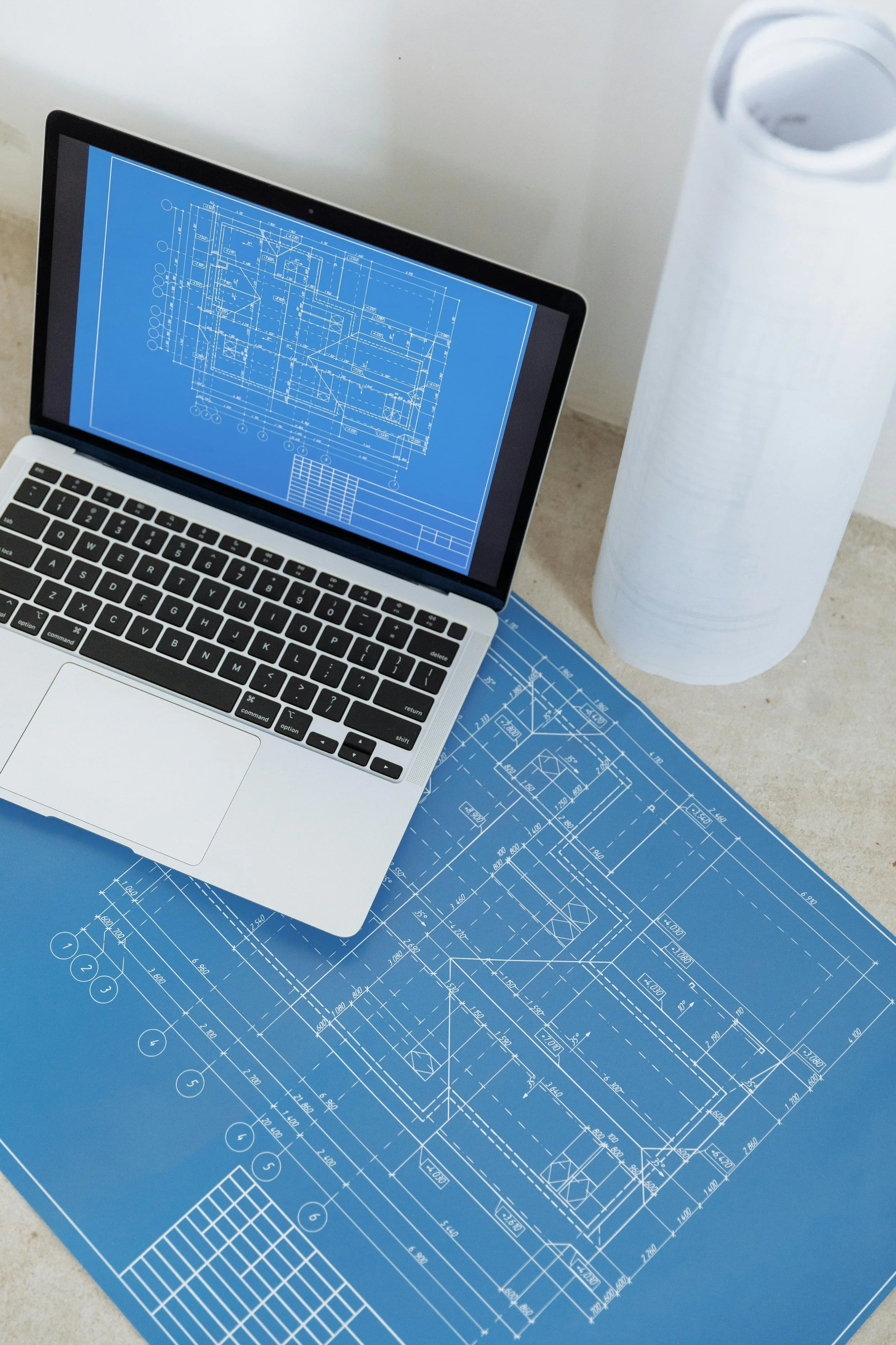 A laptop displaying a technical architectural blueprint, placed on top of a larger blueprint sheet surrounded by other blueprint sheets, and a white cylindrical object, possibly a water bottle or container.
