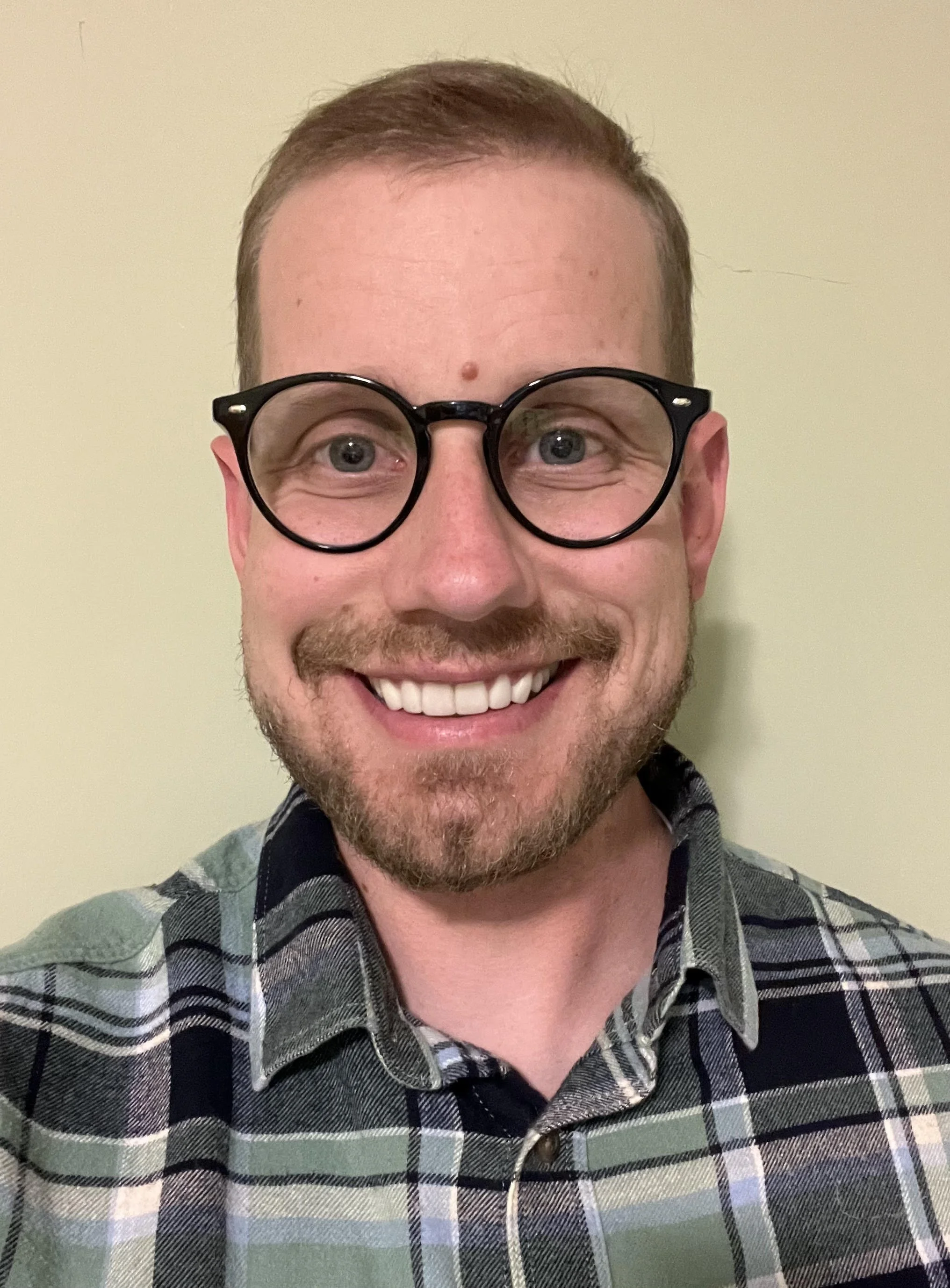 A smiling man with glasses, short light brown hair, a beard and mustache, wearing a green, gray, black, and white plaid shirt, standing in front of a plain beige wall.