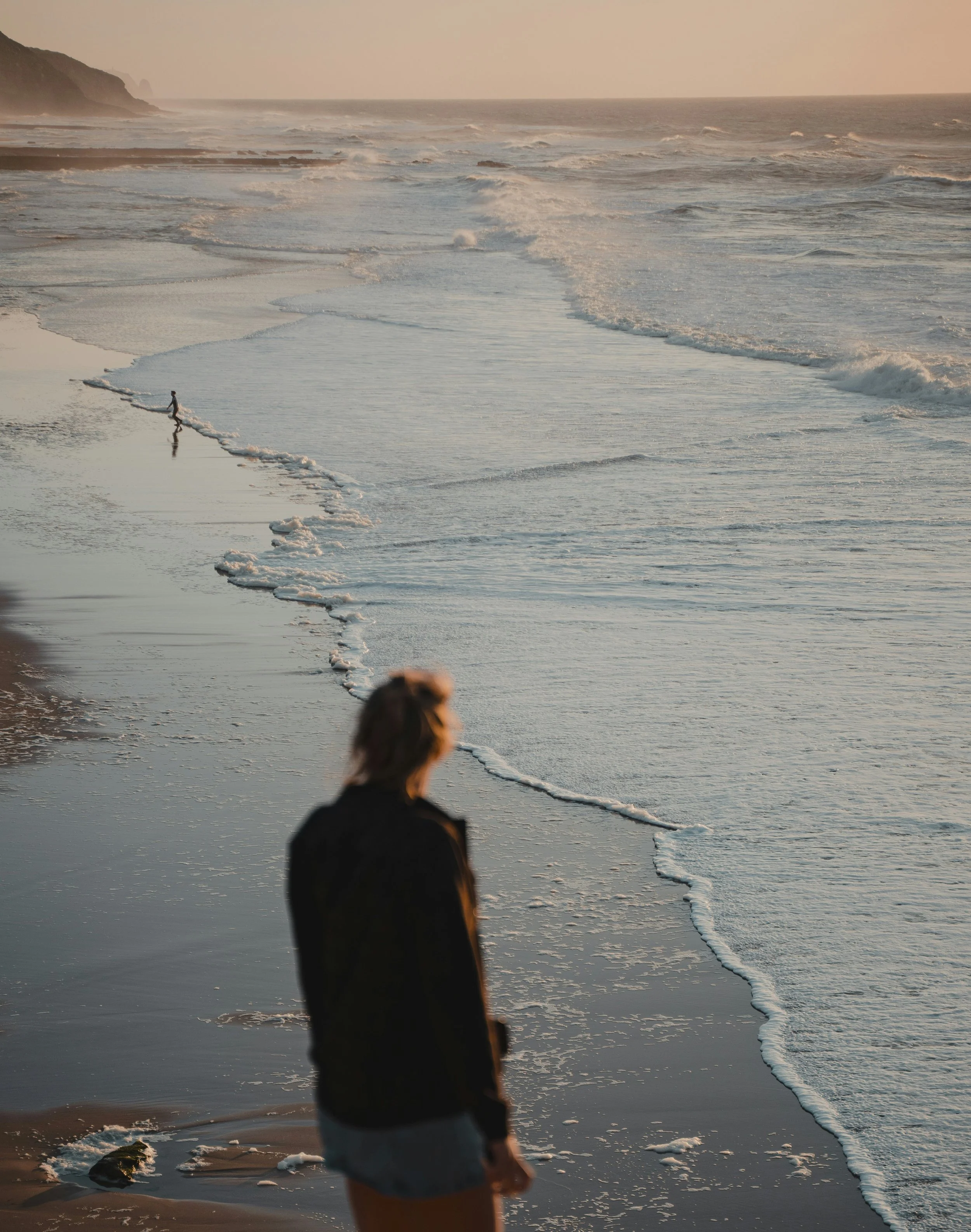 Person standing on the beach near the water's edge with waves crashing nearby, cliffs in the background, and a distant hazy horizon at sunset or sunrise.