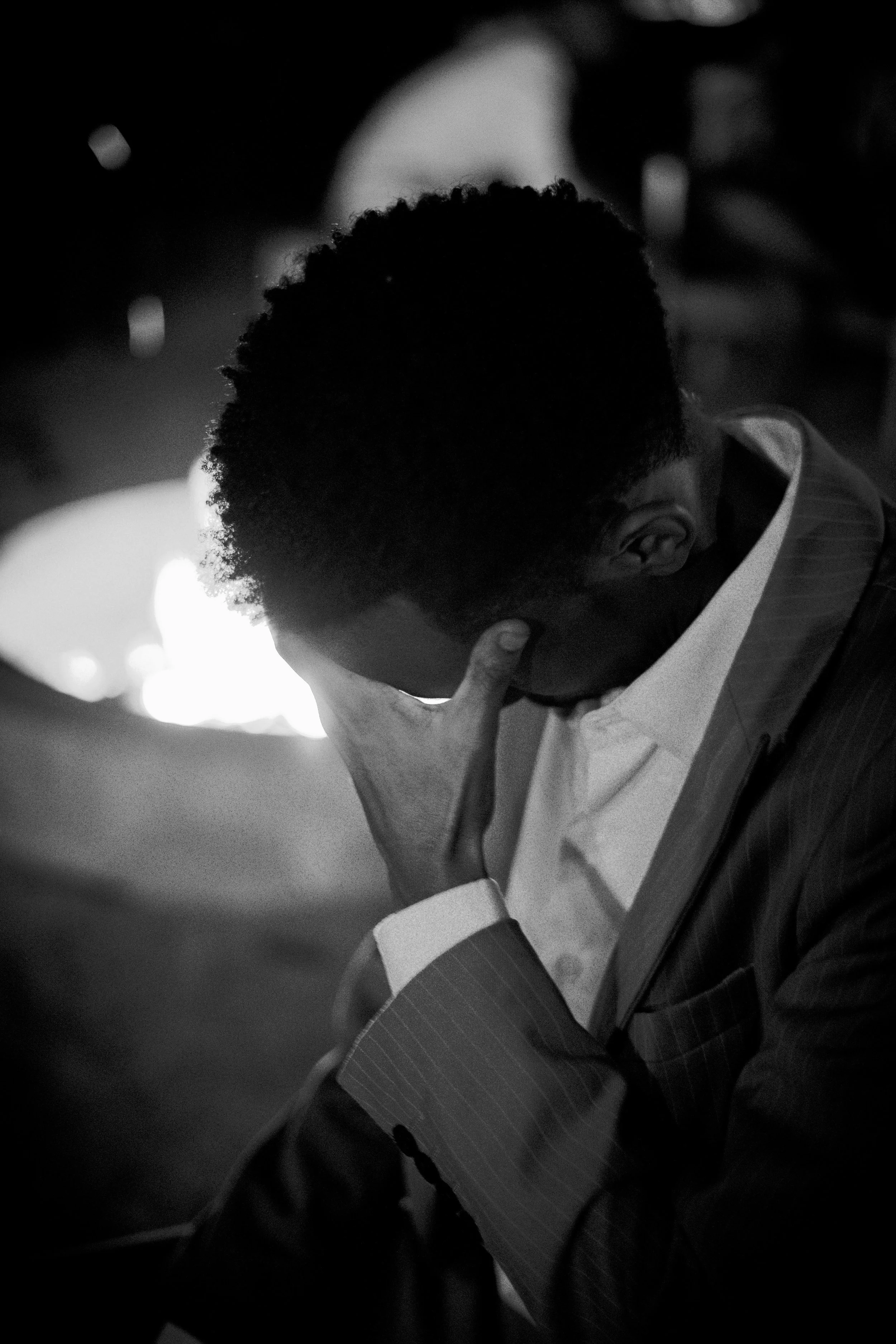 A man in a pinstripe suit and white shirt is holding his forehead with his right hand, looking down and appearing to be deep in thought or prayer. The background is dark and blurry, with some light orbs visible.