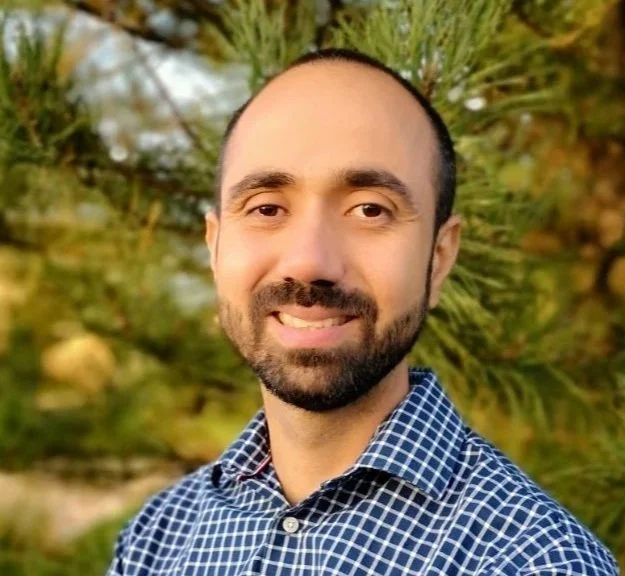 A man with a beard and short hair, smiling, wearing a blue checkered shirt, standing outdoors with blurred green trees in background.