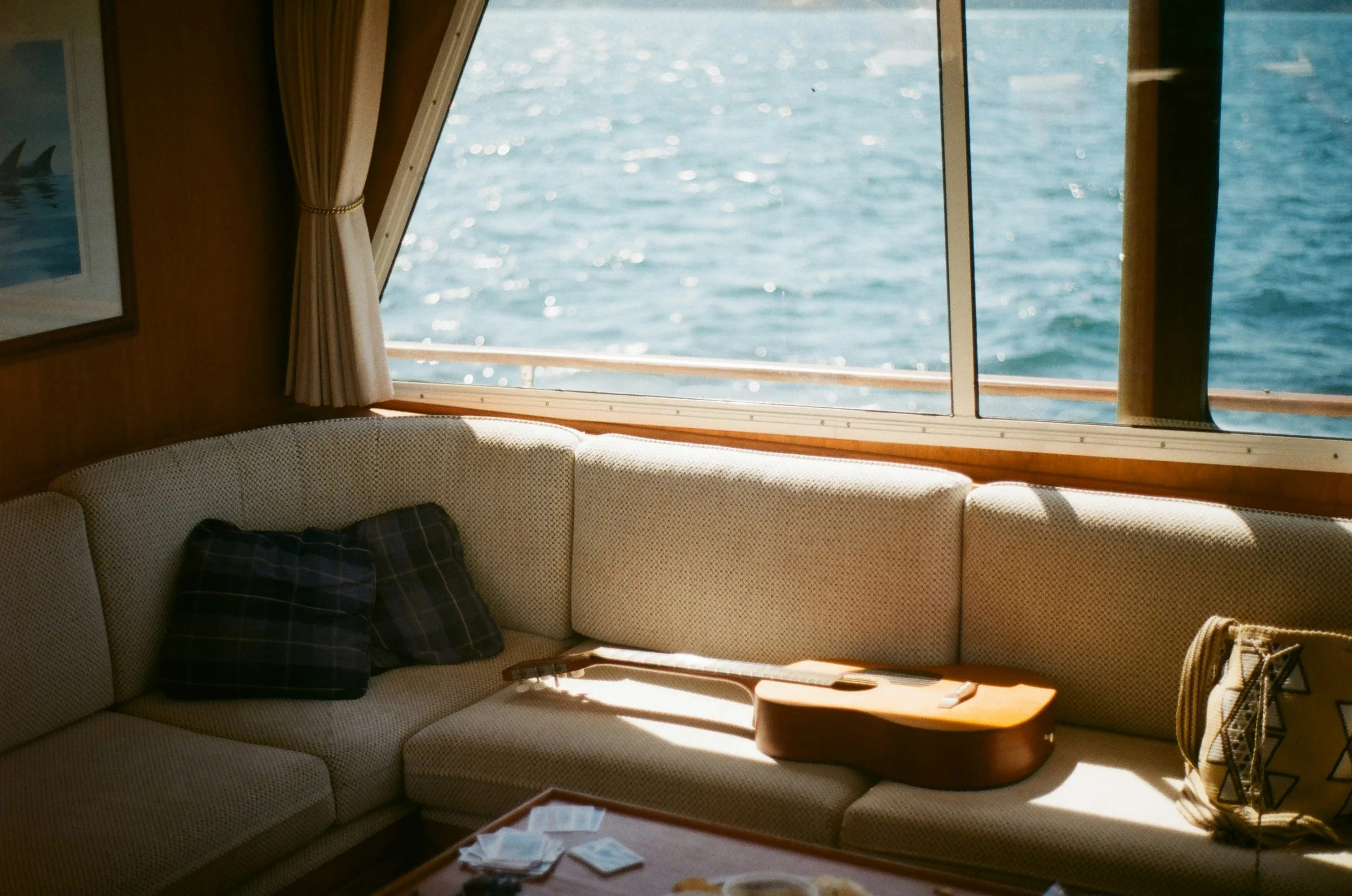 Interior of a boat or yacht cabin with a beige sectional sofa, a window overlooking water, and a curtain tied back. There are cushions on the sofa, and a small coffee table with some papers and objects. Outside, the water sparkles in sunlight.