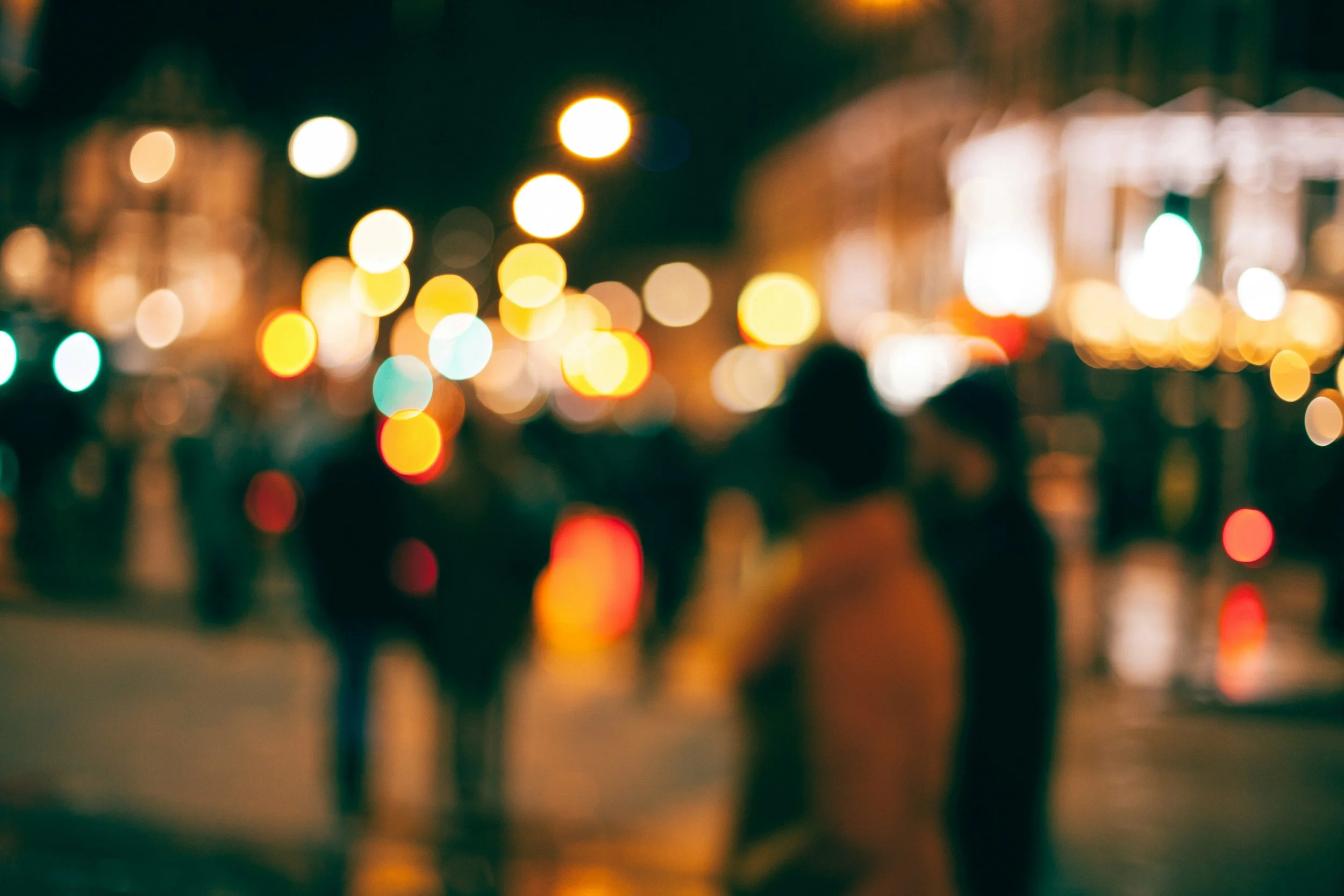 Blurred city street scene at night with colorful bokeh lights and indistinct silhouettes of people walking.